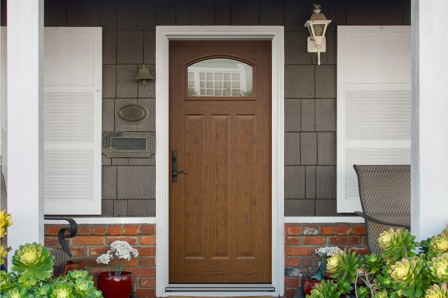 Composite wood front door with a white frame from the exterior view of a home with gray siding and a short brick facade along the bottom.