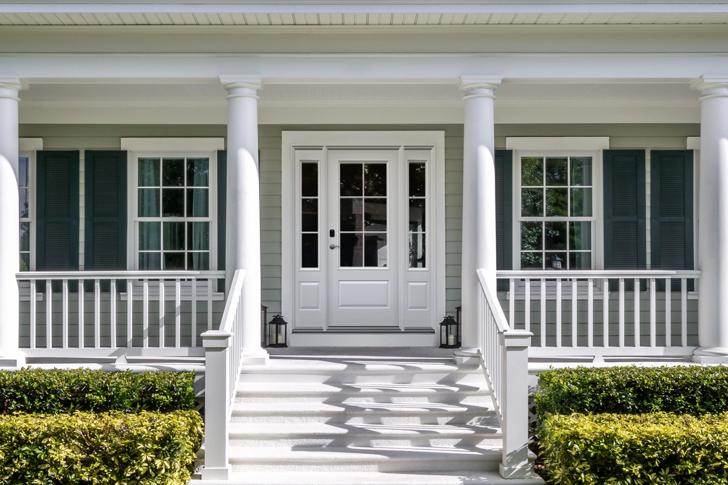 Exterior view of a white entry door with a glass panel in the middle installed on a colonial-style home with a large porch.
