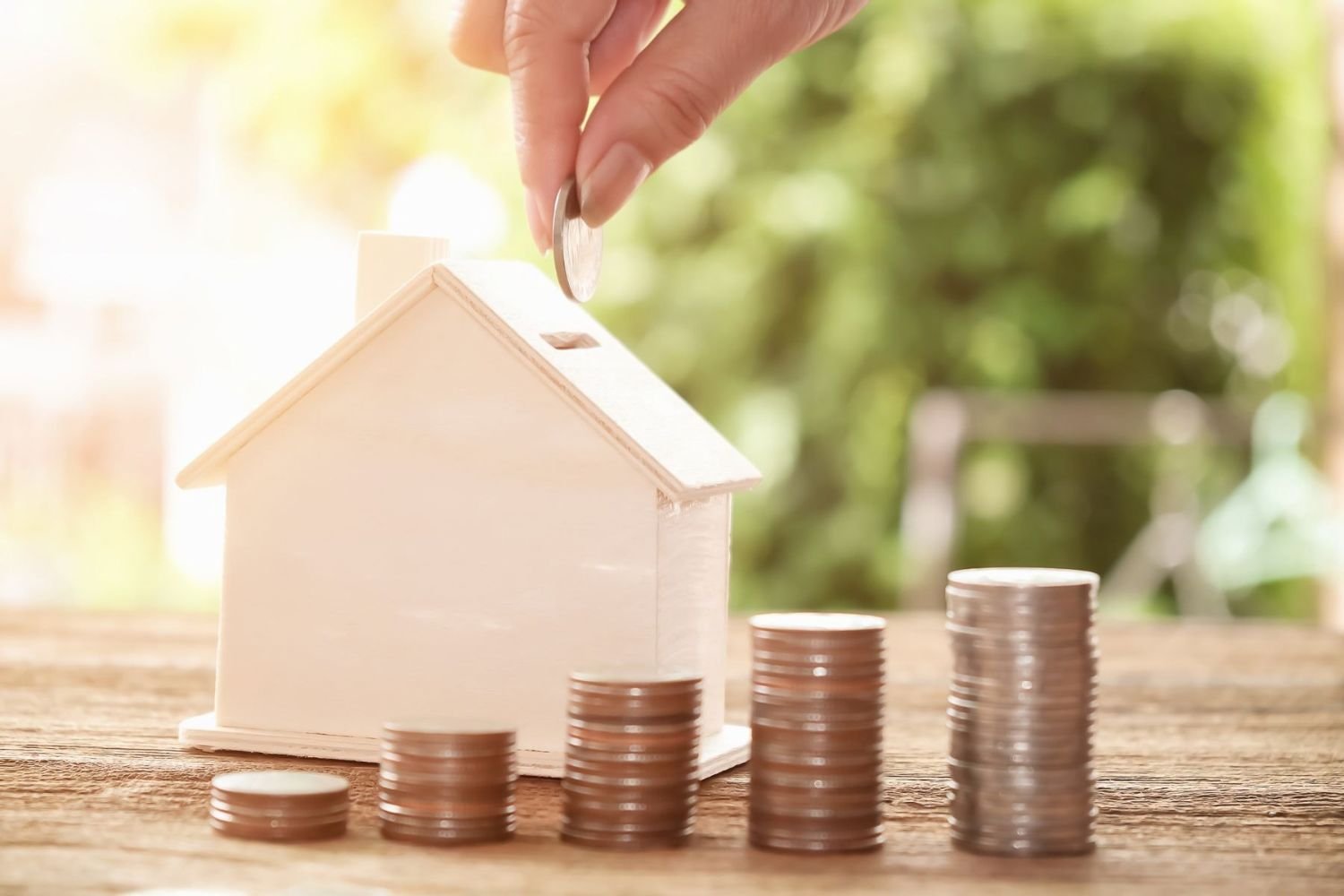 5 stacks of coins that get increasingly large from left to right sitting in front of a home shaped piggy bank above which a person's hand can be seen putting a coin into the slot.