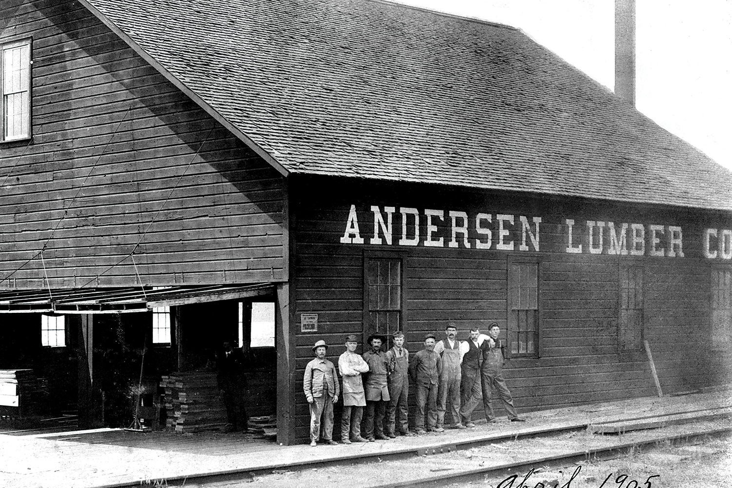 Historic photograph of the original Andersen Lumber Company building with workers standing outside the lumber mill, representing the early beginnings of Andersen Windows