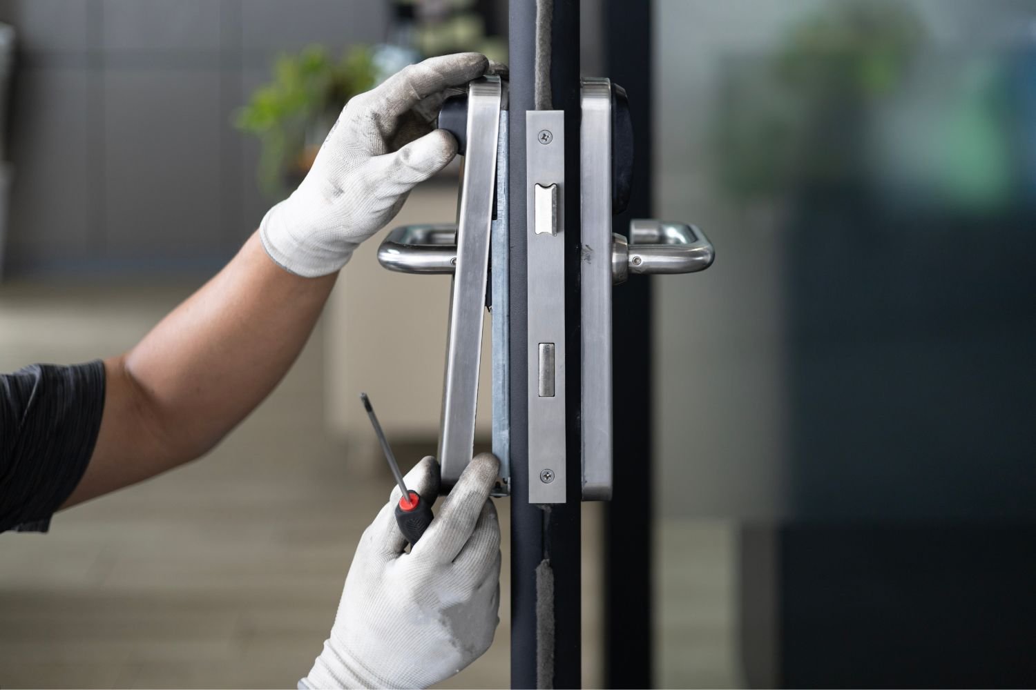Gloved hands use a screwdriver to adjust or install a metal lock mechanism on an entry door.