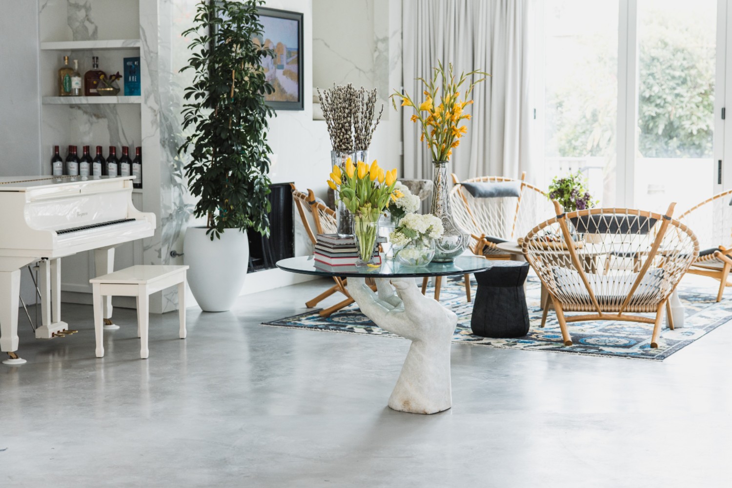 A bright, modern living room featuring a white grand piano, a sculptural hand‑shaped glass‑top table with vases of yellow and white flowers, woven lounge chairs, a blue patterned rug, and large windows with sheer curtains letting in natural light.