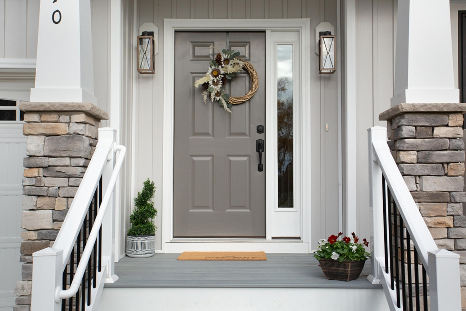 A beige/gray front door on an off-white house with a autumn-themed wreath hung in the middle. There are two white columns with stone bases framing the stairs that lead up to the door.