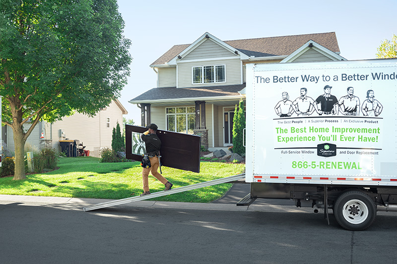 Door installer carrying a new front door out of a delivery truck to install in the two-story house that the truck is parked in front of.