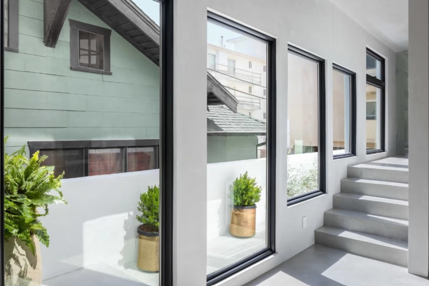 Modern interior hallway with large black-framed windows, concrete stairs, and potted plants, overlooking a neighboring building.