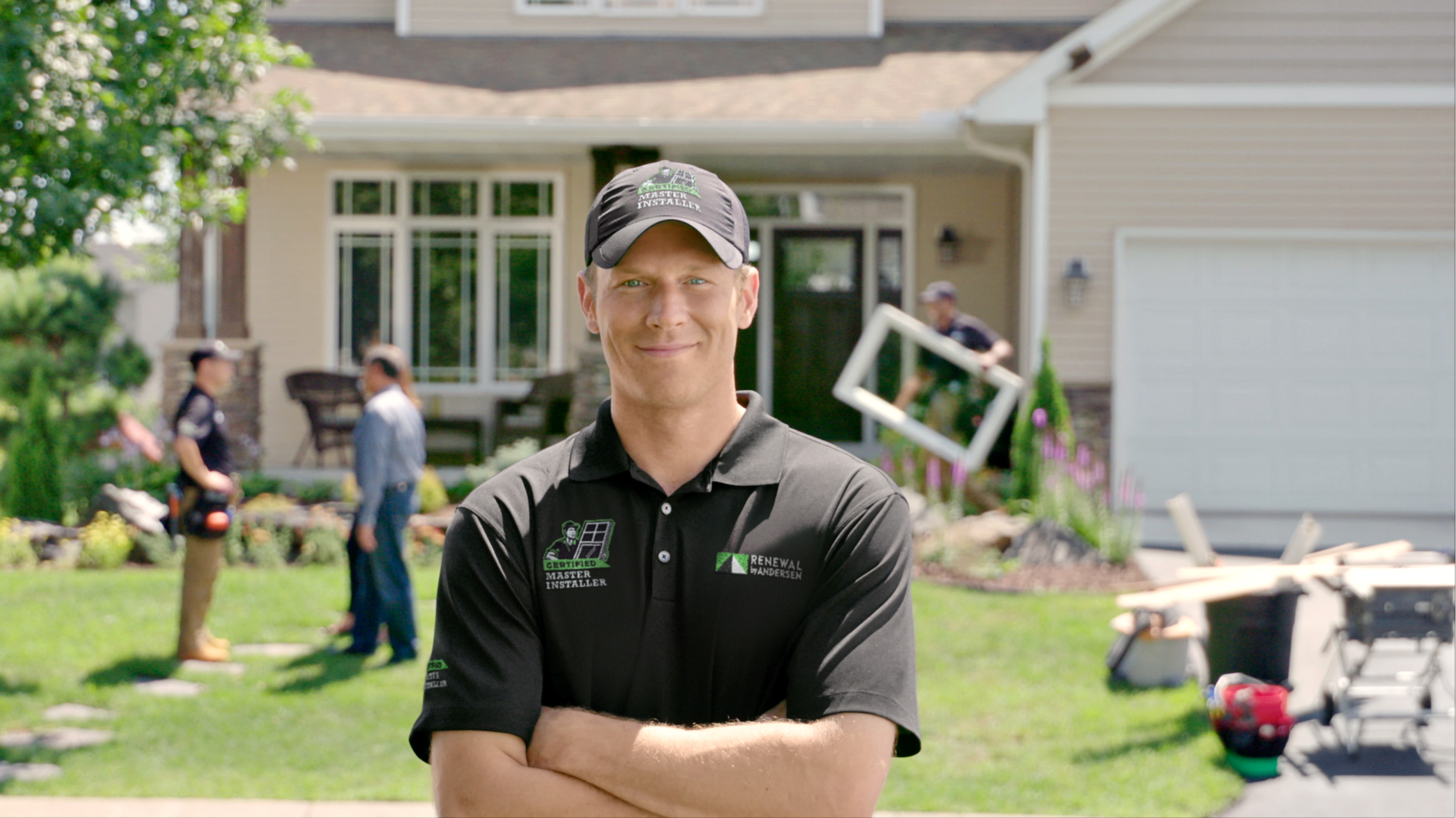 A man wearing a black shirt and hat stands in front of a house, smiling at the camera.