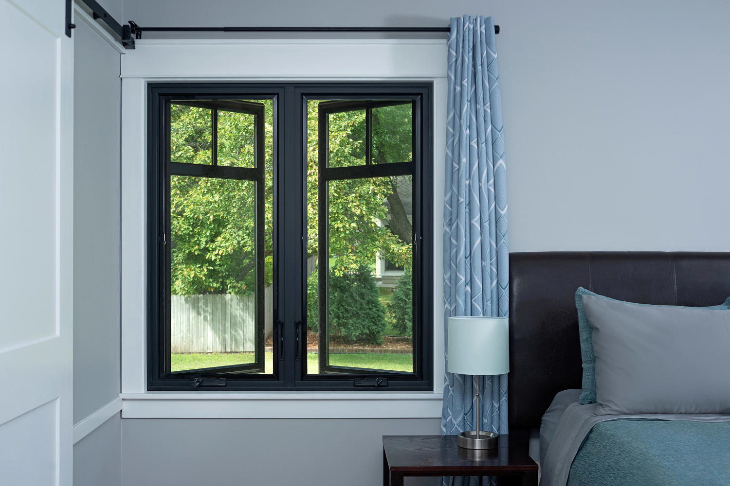 Black casement windows that are open in a bedroom. They are just to the left of the bed's headboard.