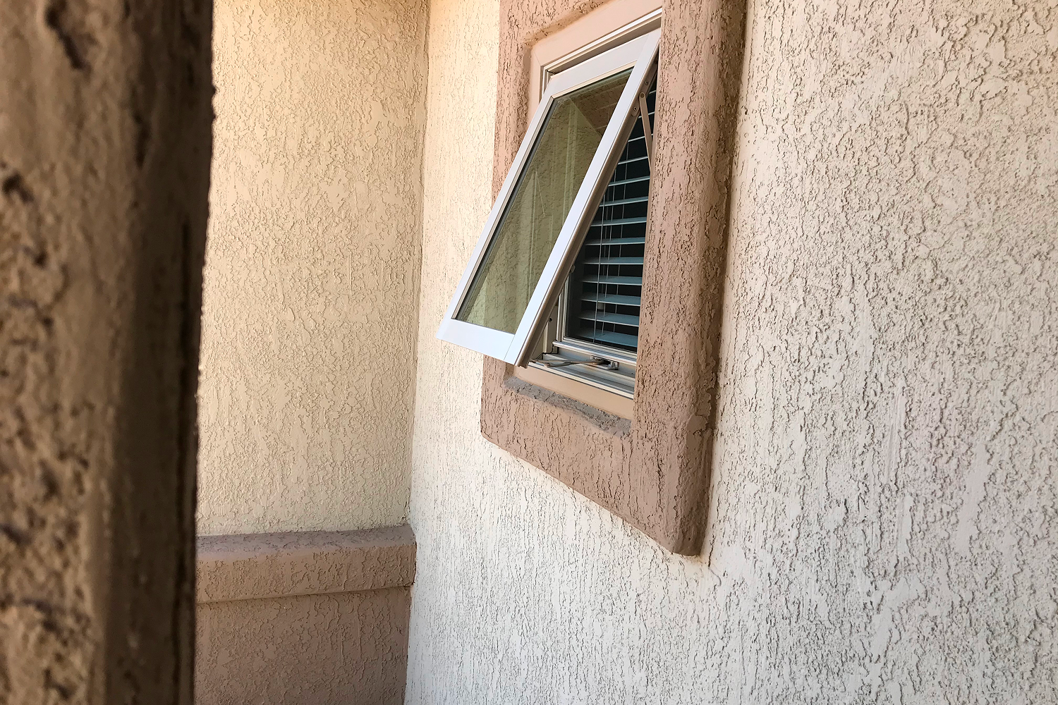An awning window in a basement shown from the outside of a stucco house.