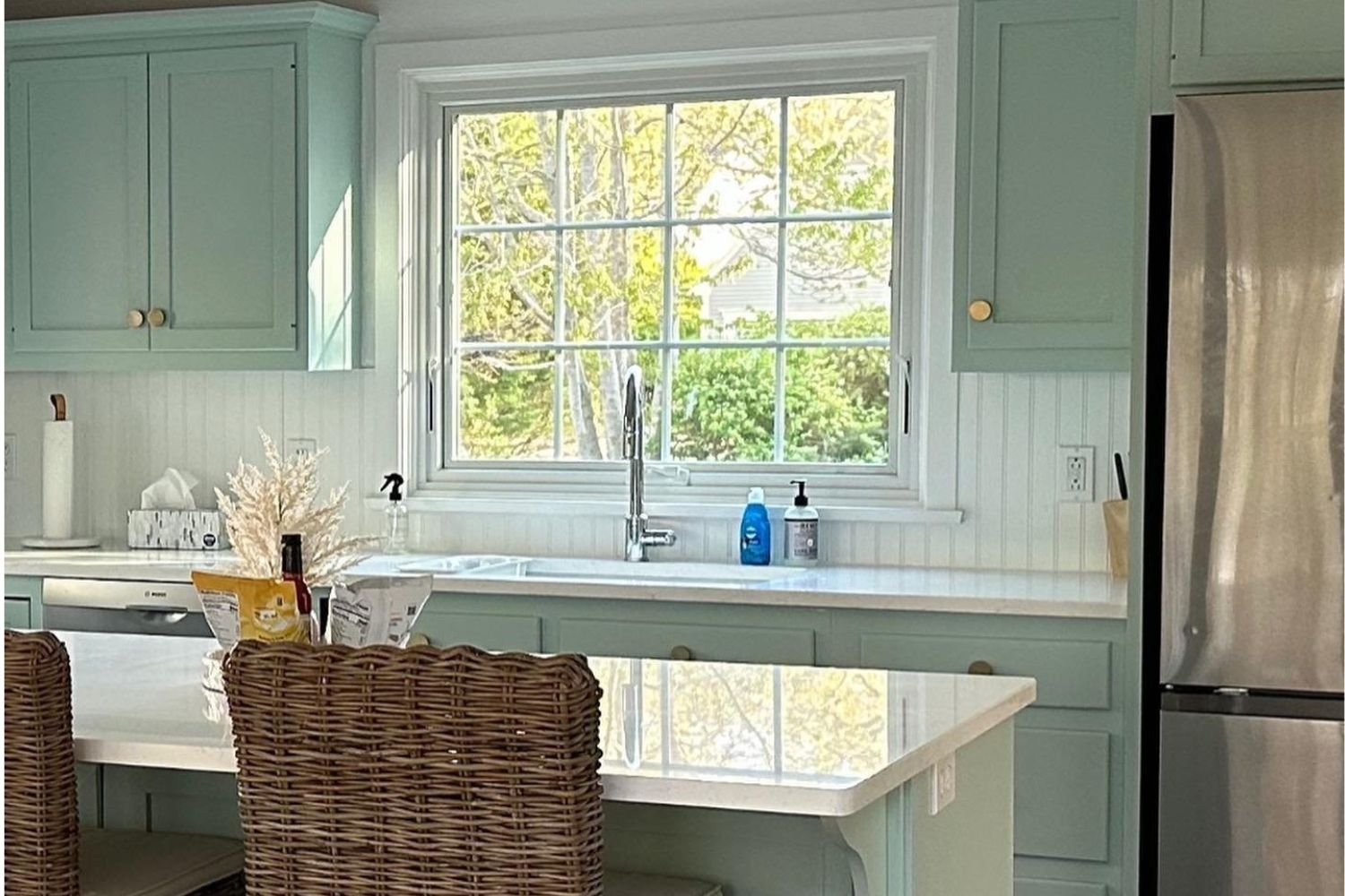 A white kitchen with blue cabinets with a large awning window behind the kitchen sink.