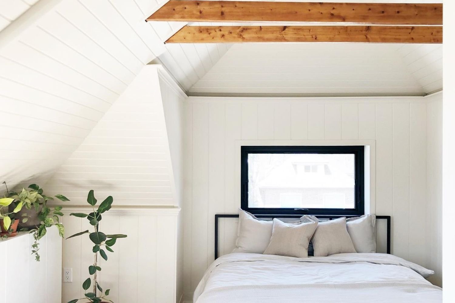 white attic bedroom with wood beams across the ceiling. There is an awning window above the headboard of the bed.