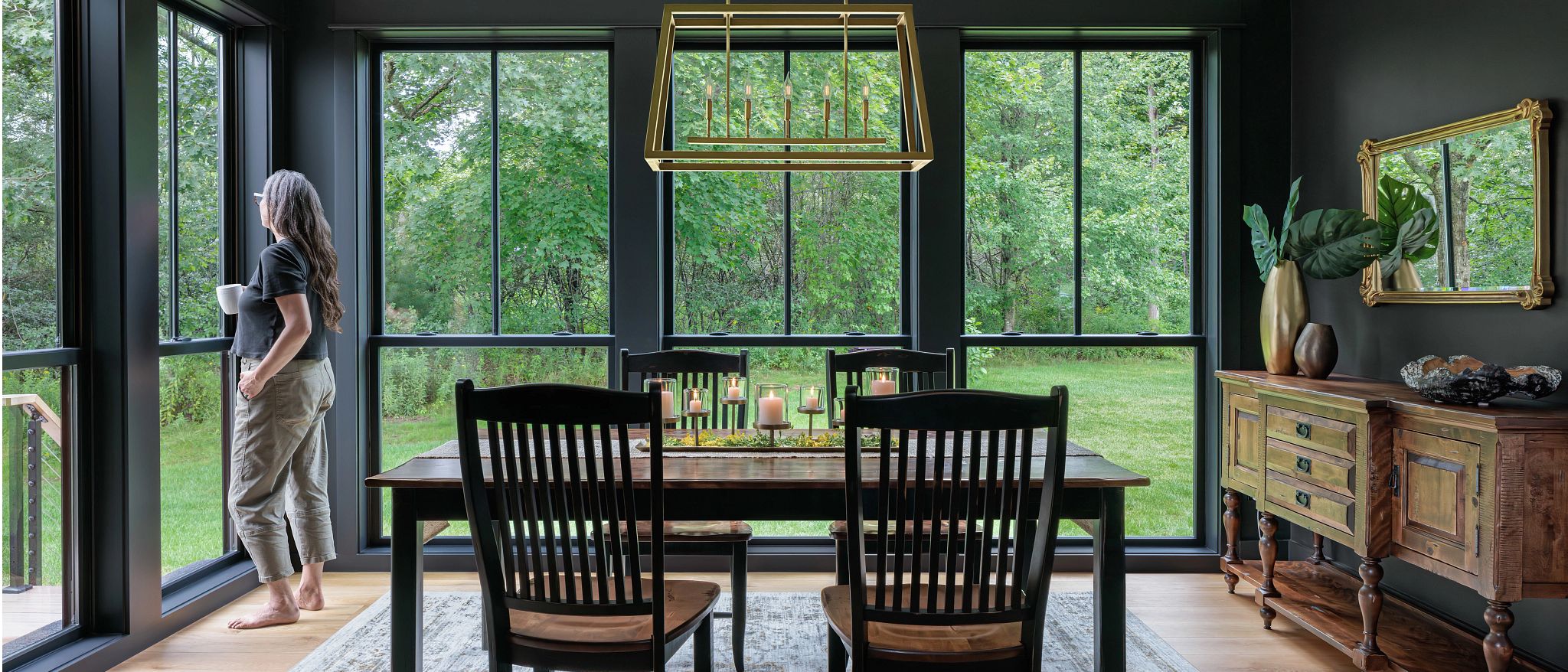 A woman stands and thinks and contemplates the fate of the world as she sips coffee. She is in a dining room with very large windows and a lovely view.