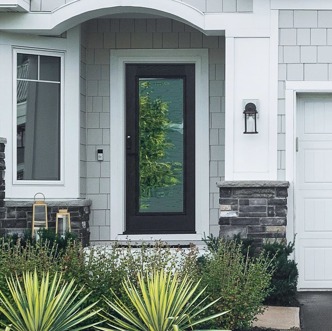 A single panel Ensemble entry door. The door is black and has full-light glass. It has white trim. You can see grey siding on the house and some spiky plants.