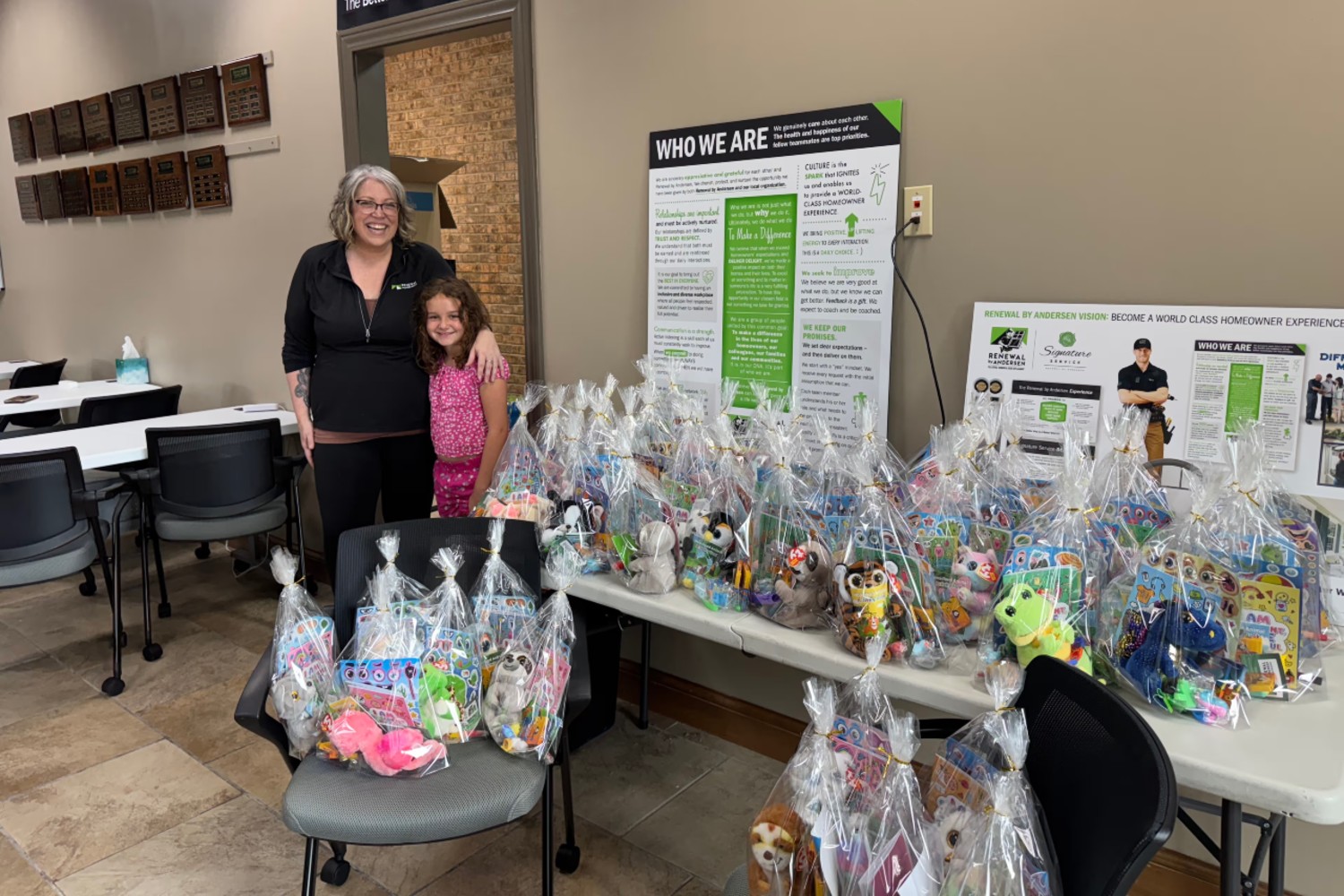 A woman and a child stand beside several tables filled with dozens of cellophane-wrapped gift bags containing toys, books, games, and stuffed animals.