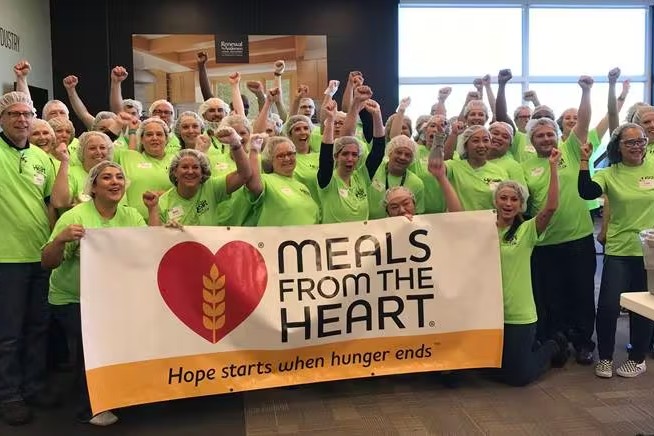 A group of about 50 Renewal by Andersen employees stand together holding a "Meals From The Heart" banner and raising their hands in excitement. 